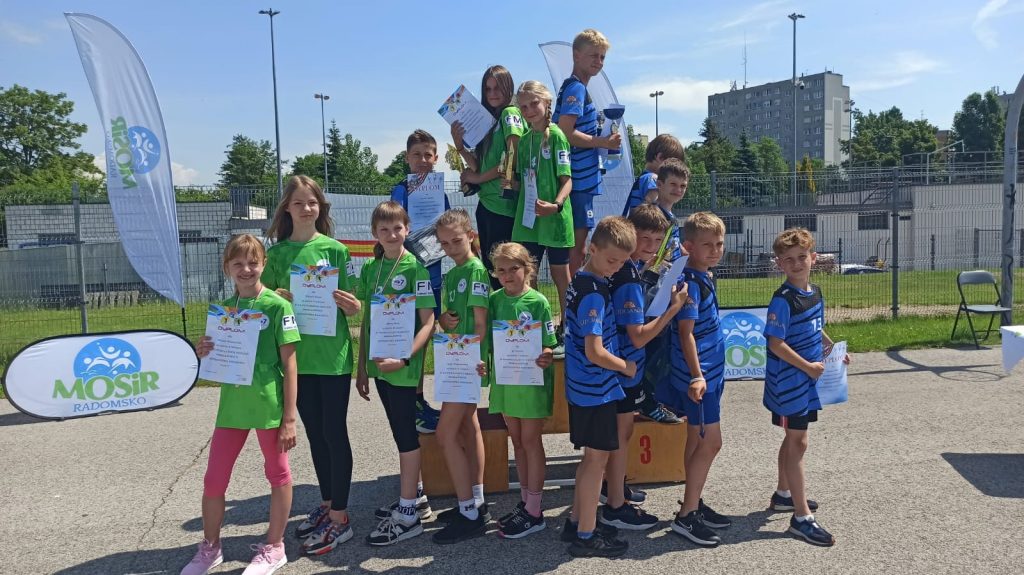 Dzieci w strojach sportowych na podium z pucharami i dyplomami.